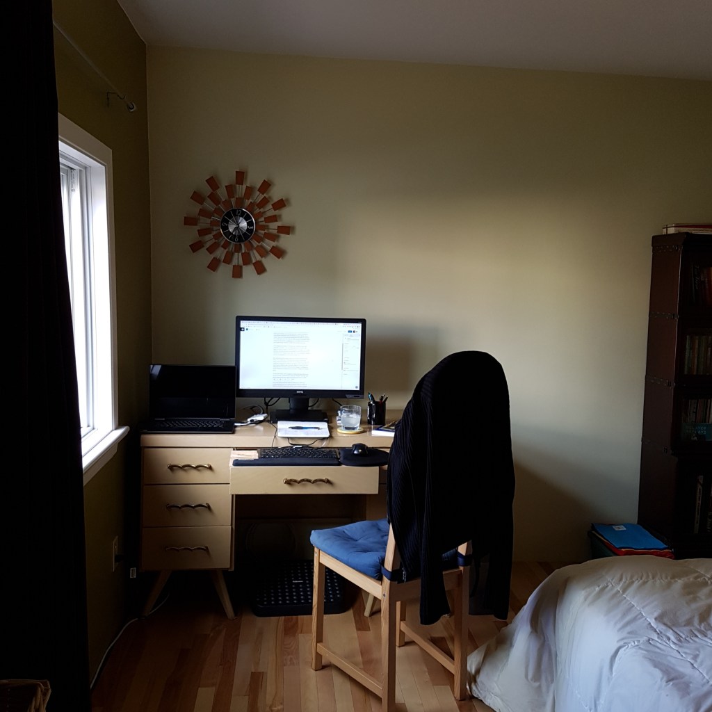 Home office. A desk with a monitor on it and a chair pulled out and slightly to the right. Lit by a window from the left.